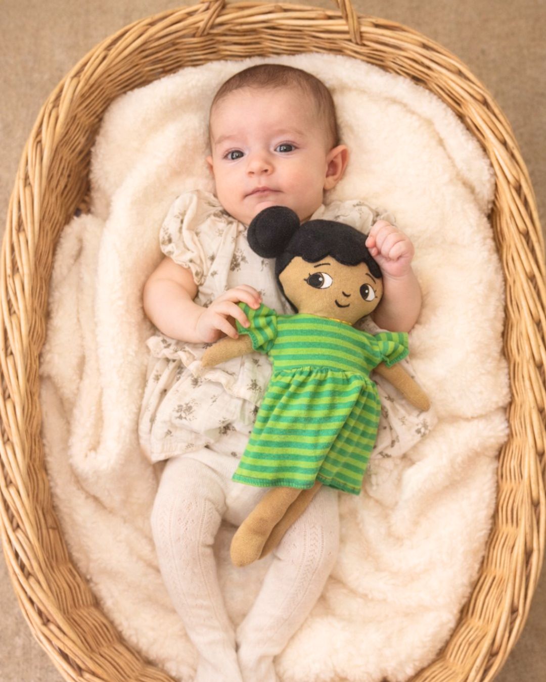 bebis ligger i korg och håller mjuk docka Amaya i grön volang klänning