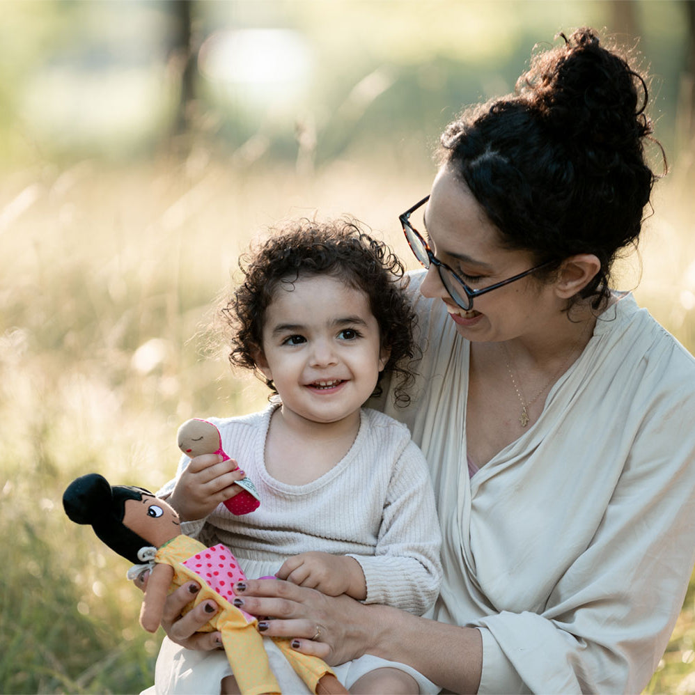 en mamma som kramar sitt barn som har en watoto arts docka i famnen. de sitter ute på gräset.