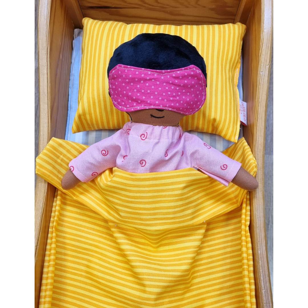 En docka som sover i docksäng, dockan bär en sovmask och har pyjamas på sig