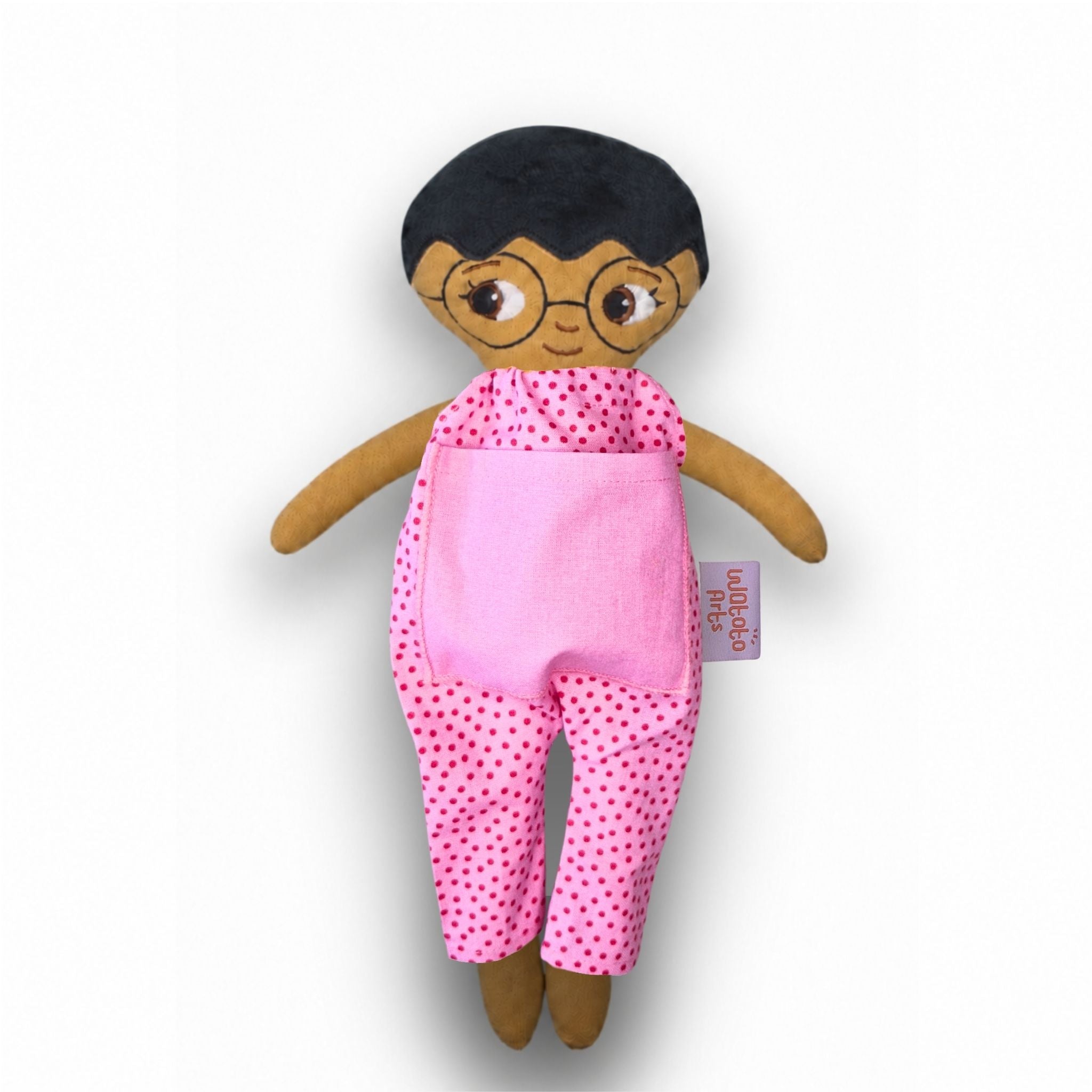 Mjuk tygdocka med kort svart hår, runda glasögon och rosa prickig jumpsuit med rosa ficka.