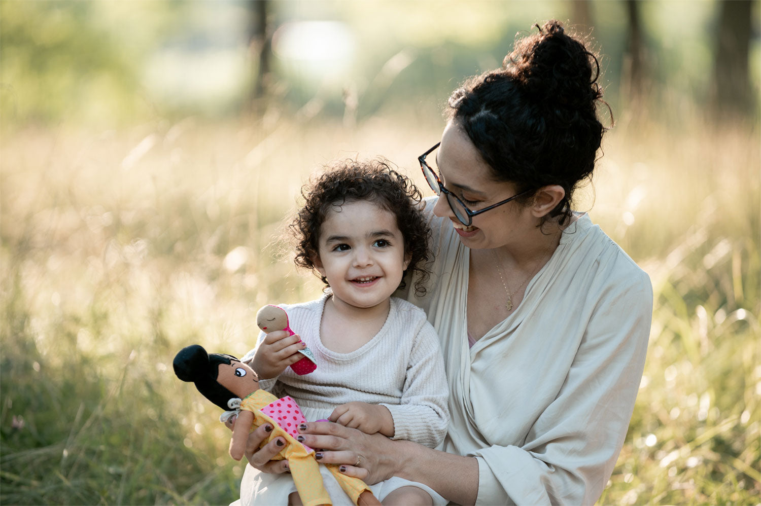 en mamma som kramar sitt barn som har en watoto arts docka i famnen. de sitter ute på gräset. 